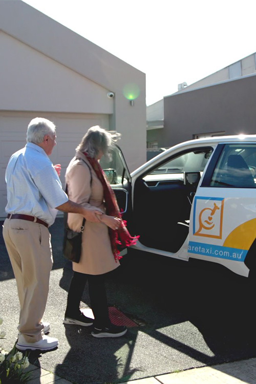 A gentleman with grey hair, wearing a blue shirt and beige trousers, helps a woman with dark hair, wearing a coat and a red scarf, into the front passenger seat of a Care Taxi car.